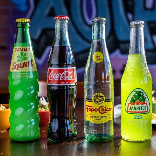 Four Mexican soda bottles, including Squirt, Coca-Cola, Topo Chico, and Jarritos Lime, on a wooden table.