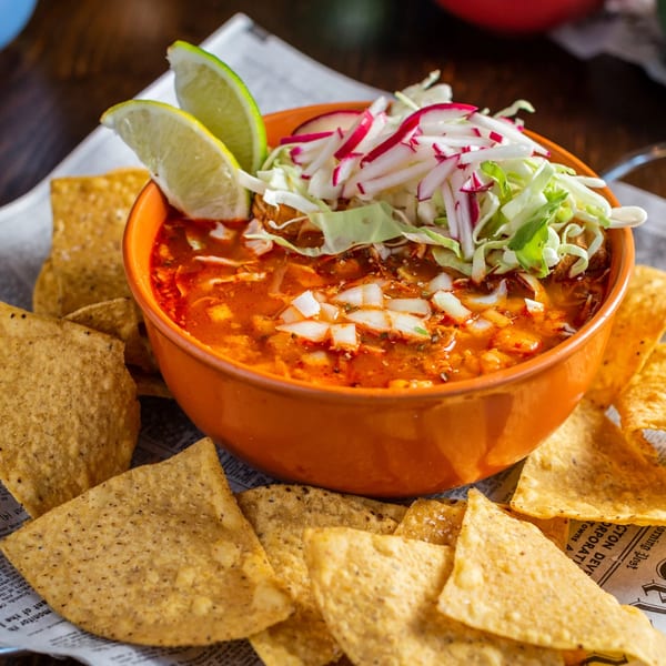 A vibrant orange bowl of Mexican pozole soup with lime, cabbage, and radishes, surrounded by tortilla chips.
