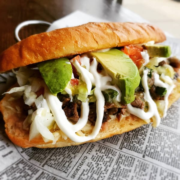 A Chicago Taco Authority Mexican torta with steak, avocado, lettuce, tomato, and crema on newspaper.