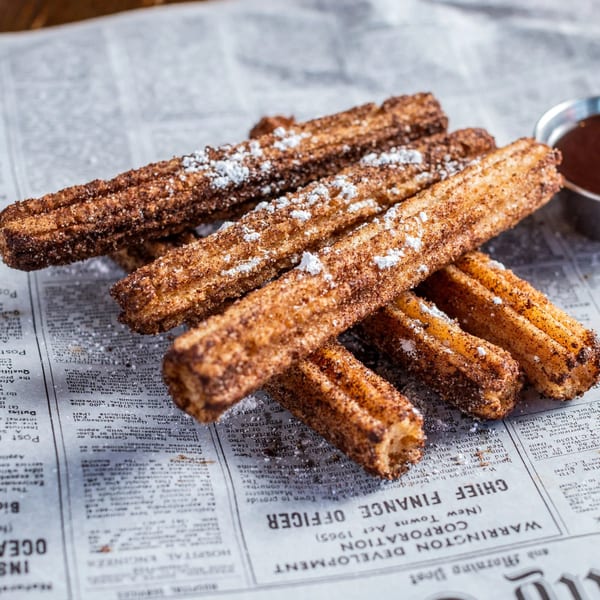 Churros dusted with cinnamon sugar and powdered sugar, served on newspaper with chocolate dipping sauce.